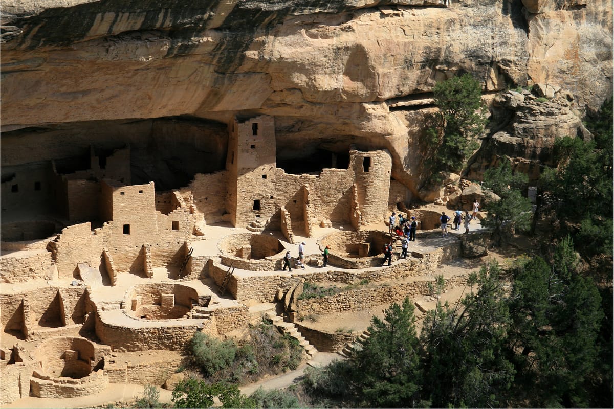 The masonry towers and stacked rooms of Cliff Palace, with visitors giving a sense of the dwelling's scale beneath the alcove's roof.