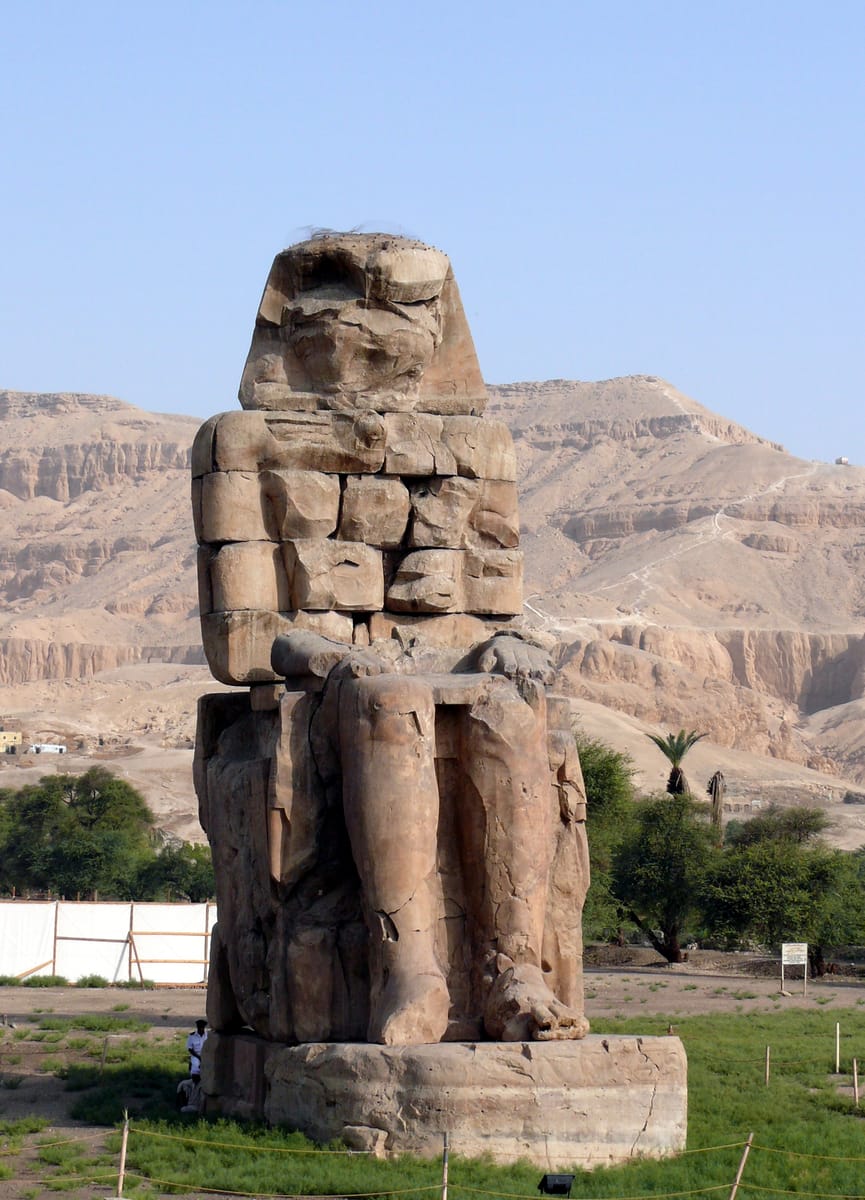 A single colossus seen from below. The faces and much of the surface detail are lost to nearly 3,400 years of weathering, flooding, earthquakes, and human damage, but the enthroned pose and monumental scale remain.