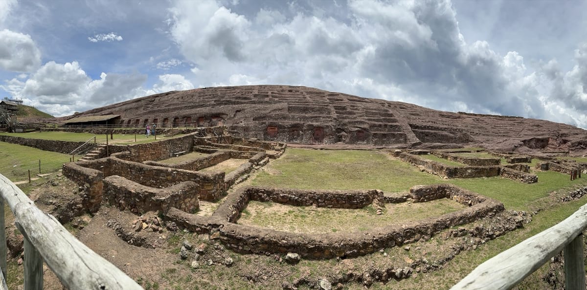 Panorama of the site: the carved sandstone hill with its rows of niches rising behind the excavated stone foundations of buildings at its base.