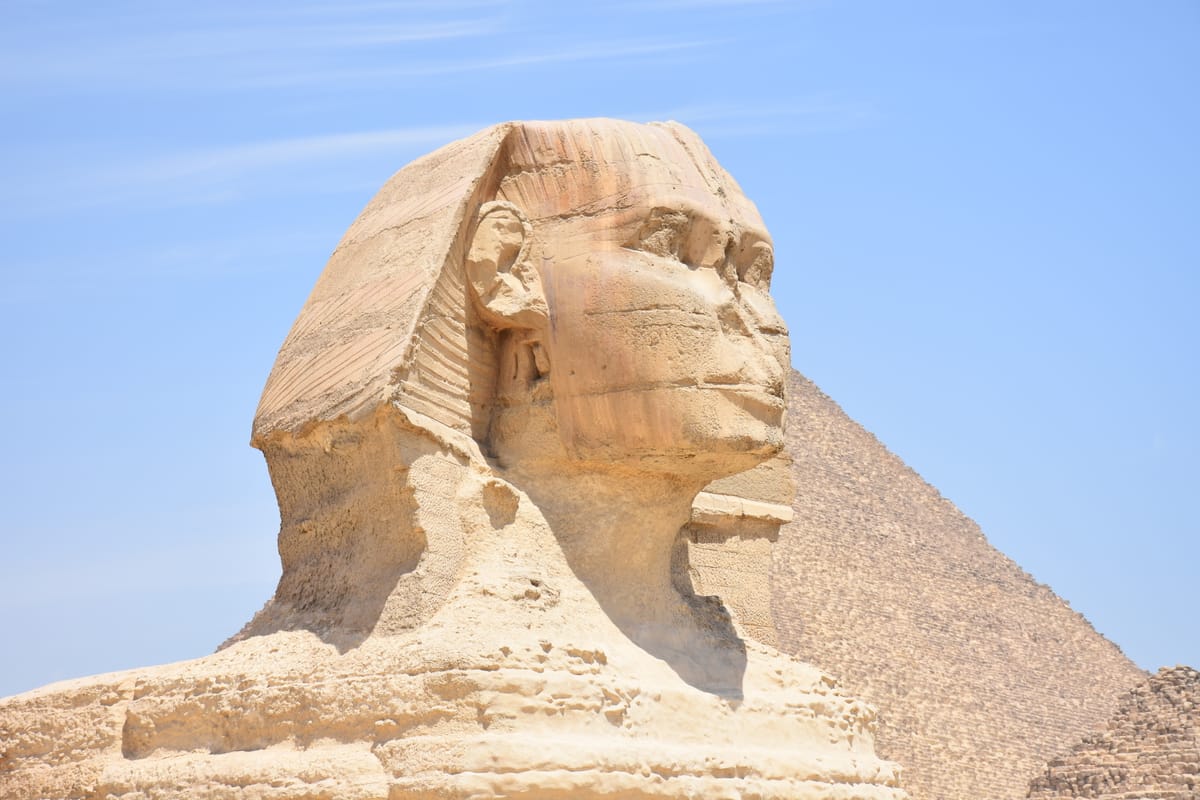 The head in close view. The nemes headcloth frames a face that has lost its nose — deliberately chiselled off — and the ceremonial beard that once projected from the chin.