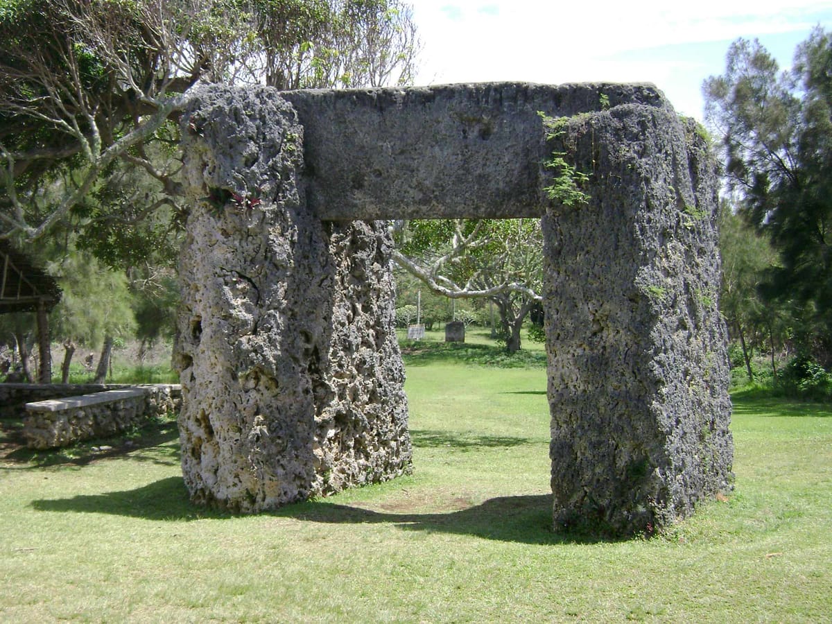 The trilithon viewed through its own opening, with the 'esi maka fa'akinanga — the upright 'stone to lean against', traditionally the king's back-guarded seat — visible in the distance beyond.