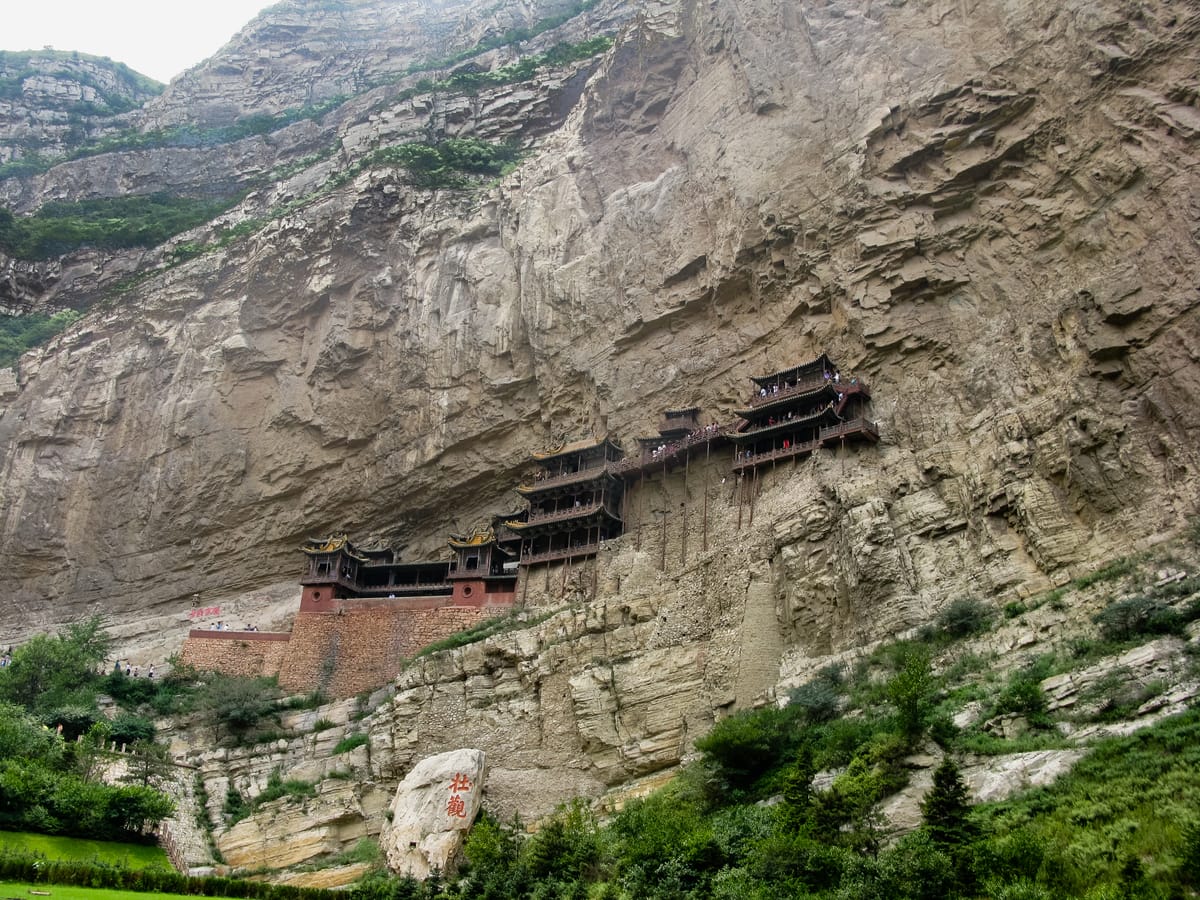 The monastery seen from the gorge floor beneath a towering overhang; the boulder in the foreground carries the characters 壮观 ('magnificent'), a phrase linked by legend to the Tang poet Li Bai.