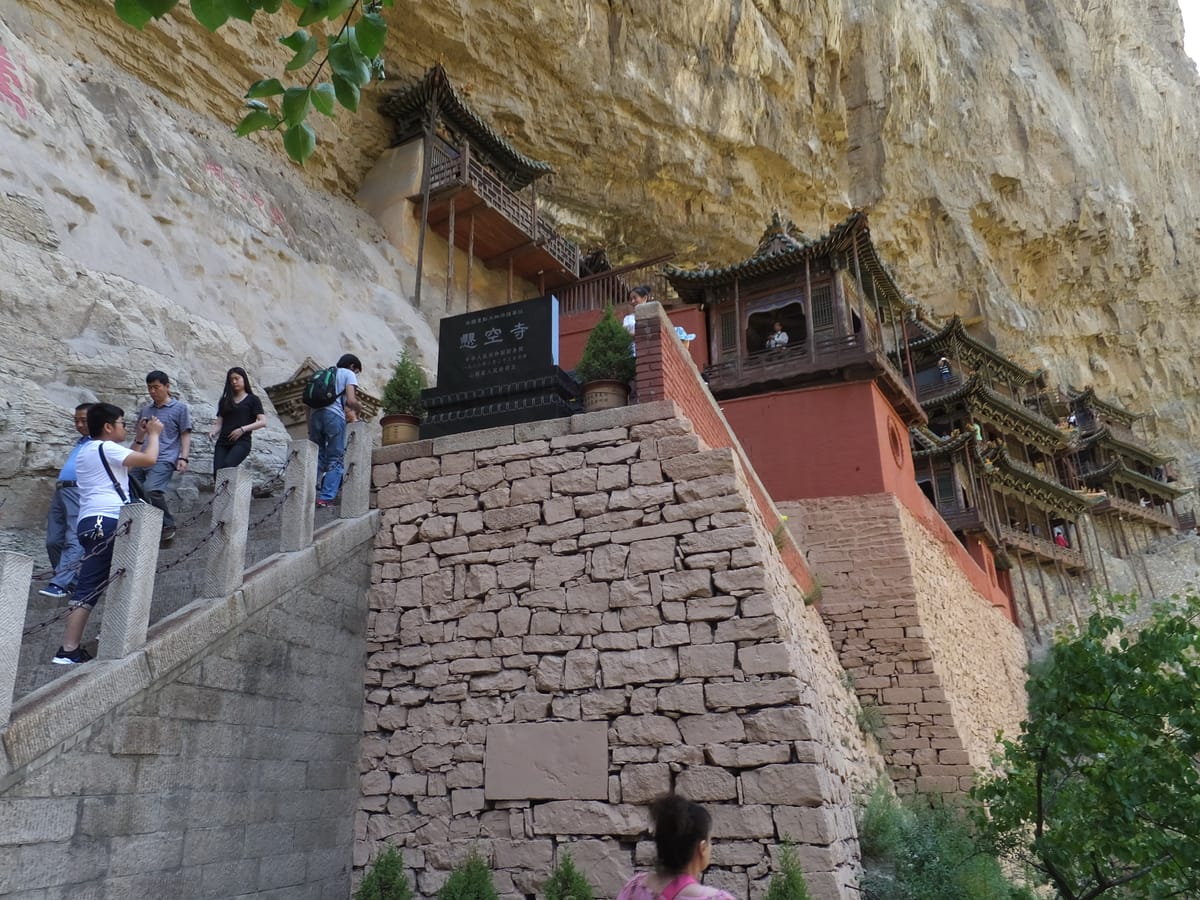A closer view from the approach stairway, past the stone stele reading 悬空寺 ('Hanging Temple'), showing the thin support poles and the beams anchoring the halls to the cliff.