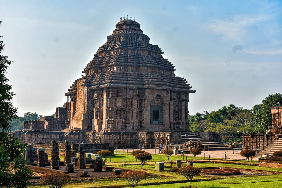 The pyramidal jagamohana seen across the ruined temple grounds, with two of the chariot wheels visible along its plinth — the sanctuary tower that once rose behind it is entirely gone.