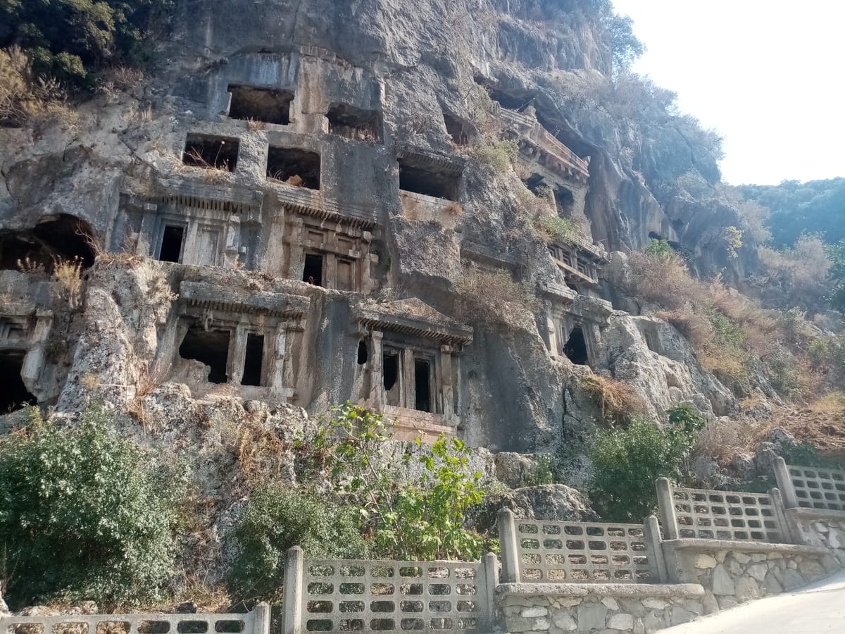 House-type tombs in the cliff above Fethiye, ancient Telmessos. The projecting rows beneath each flat roof are carved beam-ends — stone copies of the timber joists of a real Lycian house.