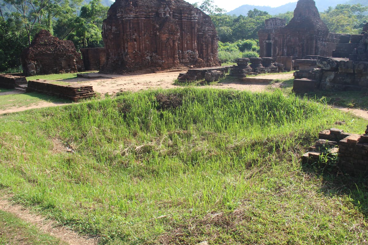 Brick towers of Mỹ Sơn beside a grassed-over depression left by American bombing in 1969; craters from the strikes are still visible across the site today.
