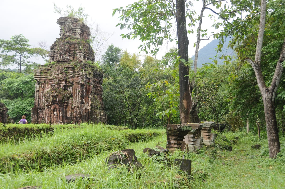 A weathered kalan rising from the forest floor of the Mỹ Sơn valley, with the surrounding hills that ring the sanctuary behind it.