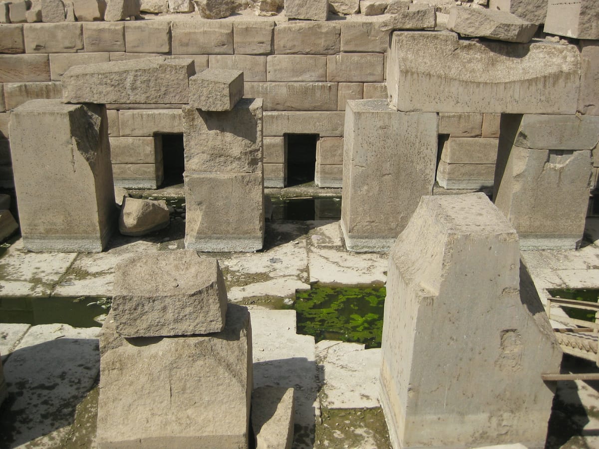 Granite piers of the central island in front of the enclosure wall, where a row of small doorless cells is cut into the surrounding masonry — the 'chambers' that ring the hall. Standing water still covers much of the floor.