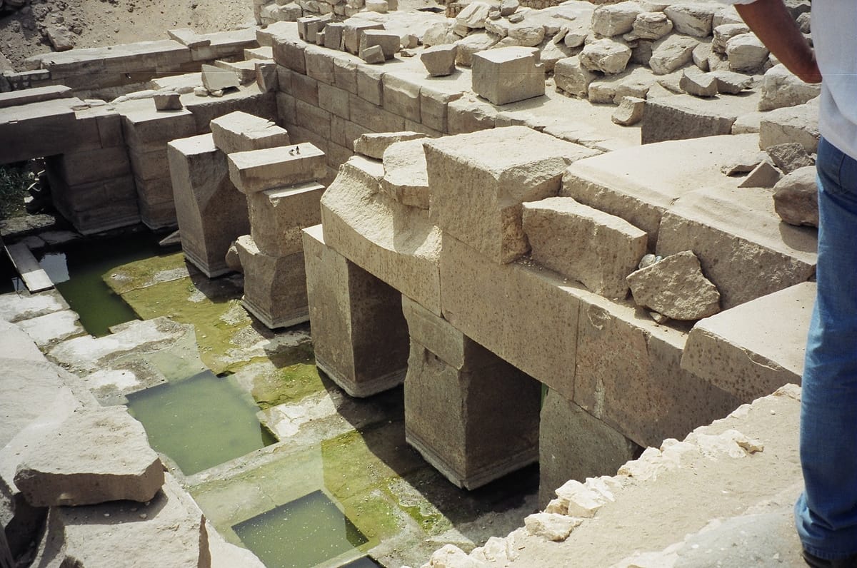 The austere post-and-lintel megalithic construction at close range — huge dressed granite piers carrying plain architraves, with water standing in the channels around the central platform. A visitor at the upper right gives a sense of scale.
