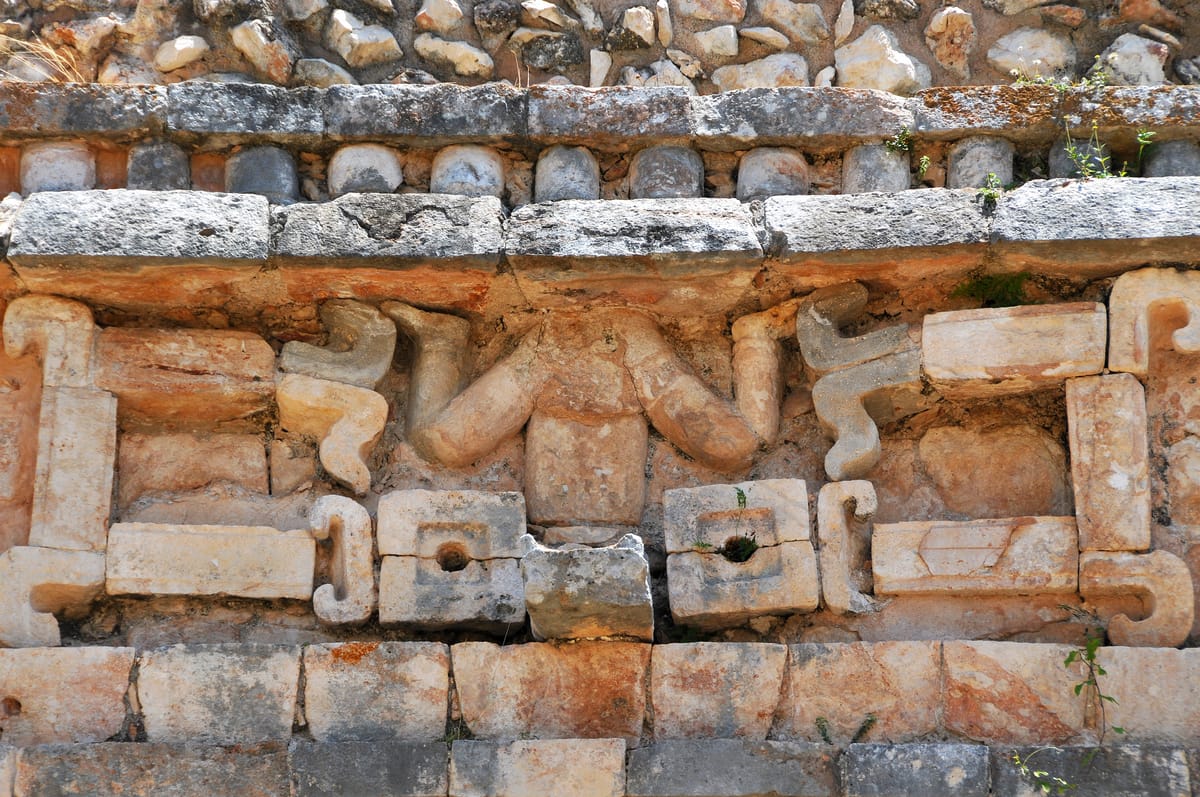 A 'descending god' on the second-range frieze — often identified as Ah Muzen Cab, the Maya bee deity — diving head-down between bands of stone colonnettes.