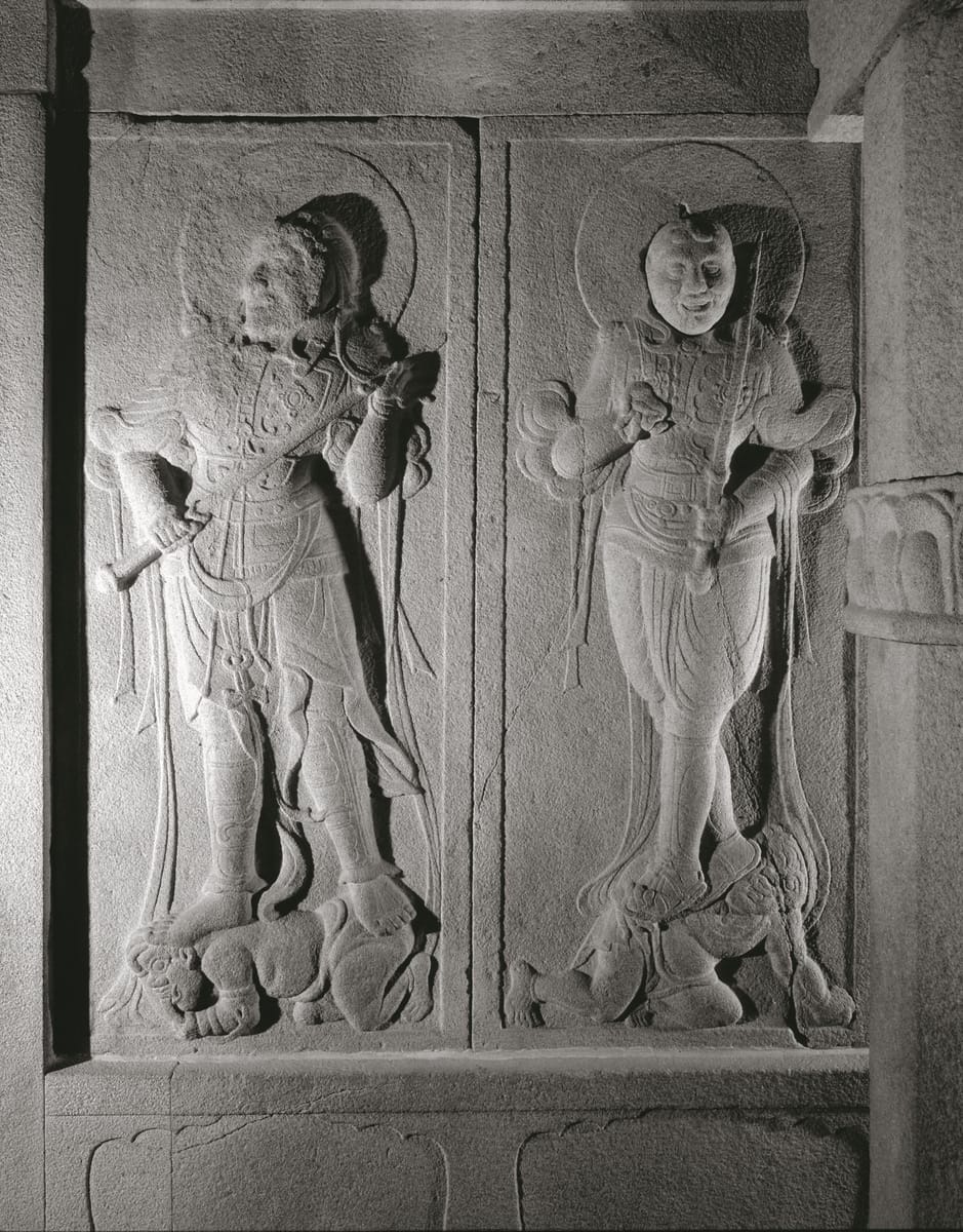 Two of the Four Heavenly Kings carved in relief on the corridor walls, each trampling a demon underfoot — the guardians a pilgrim passes before reaching the rotunda.