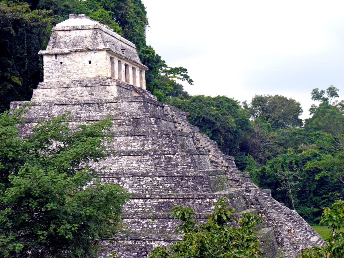 Seen obliquely, with the forested ridge behind. The heavy mass of the temple's walls is what a corbel vault demands; the openwork roof crest above lightens it.