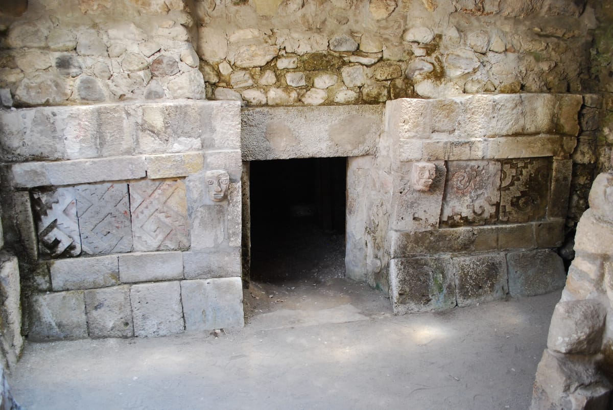 The facade of the Triple Tomb below Patio 4. Stepped meander panels — xicalcoliuhqui — still carry traces of red paint, and a small carved human head is set into the jamb on each side of the doorway, held by a long tenon buried in the wall behind it.