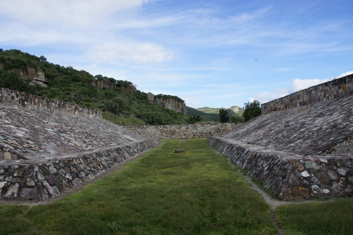 The ballcourt: an I-shaped alley about 30 metres long between sloping lateral walls of undressed river cobble, the largest court in the Valley of Oaxaca. There are no rings — Oaxacan courts never had them.